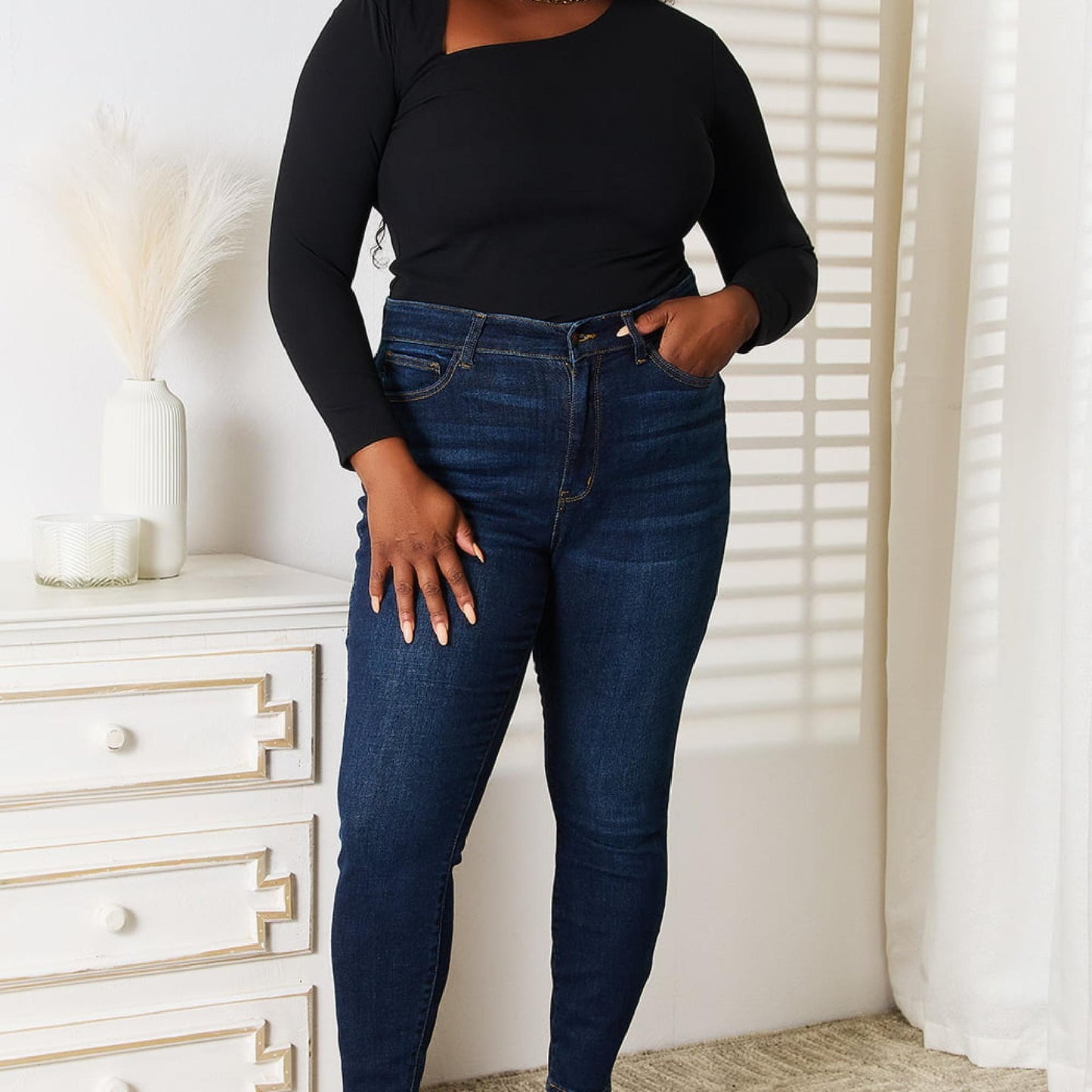 Woman wearing a black top and blue jeans standing in a room with white walls and a white dresser.