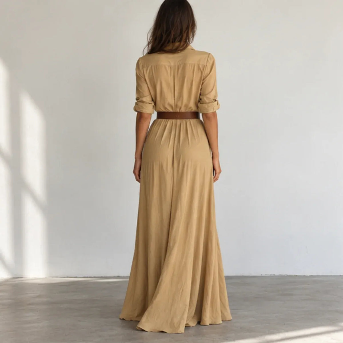 A woman viewed from behind wearing a tan maxi shirt dress with rolled sleeves and a thin brown belt at the waist, standing in a minimalist room with a white wall and concrete floor.