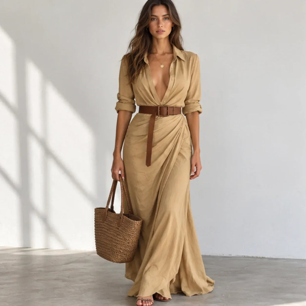 A woman models a tan maxi shirt dress with a deep V-neckline, rolled sleeves, and a matching brown belt. She is holding a woven straw tote bag and standing in a bright, minimalist room with soft window shadows.