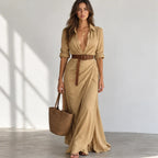 A woman models a tan maxi shirt dress with a deep V-neckline, rolled sleeves, and a matching brown belt. She is holding a woven straw tote bag and standing in a bright, minimalist room with soft window shadows.