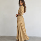 A woman modeling a beige maxi shirt dress with three-quarter sleeves and a matching belt, standing in profile against a plain white wall.