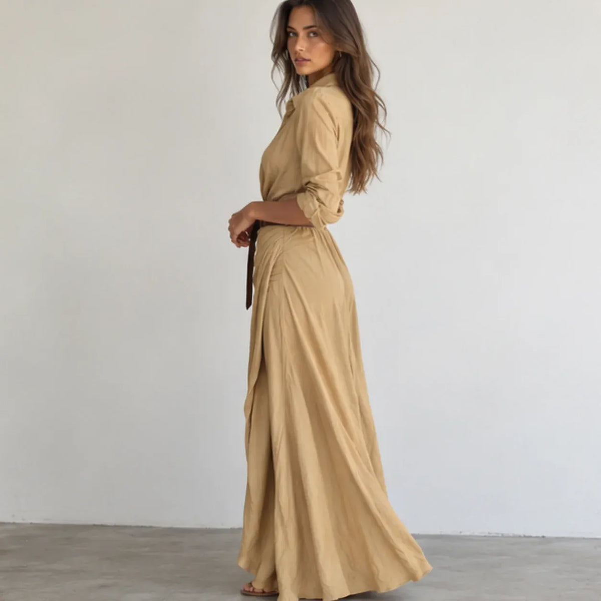 A woman modeling a beige maxi shirt dress with three-quarter sleeves and a matching belt, standing in profile against a plain white wall.