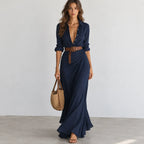 A woman models a navy blue maxi dress with a button-down shirt collar, rolled sleeves, and a brown belt. She is holding a woven straw tote bag and standing against a neutral light gray wall and concrete floor.