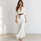 A woman models a white long-sleeve maxi shirt dress with a deep V-neckline, buttoned front, and a brown belt at the waist, set against a minimalist white studio background.
