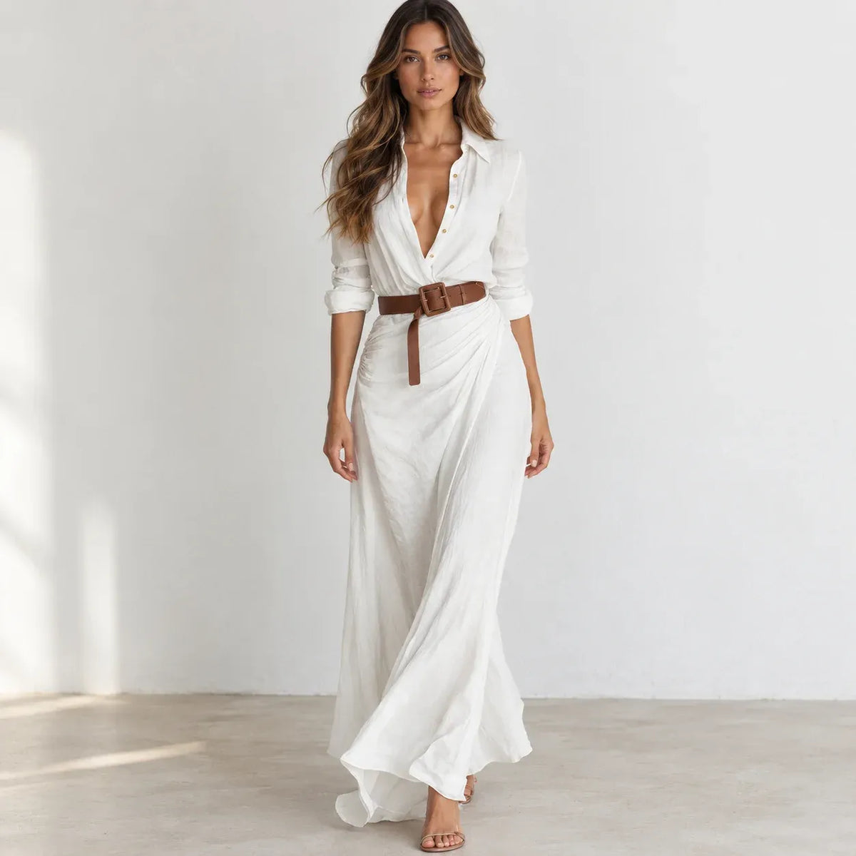 A woman models a white long-sleeve maxi shirt dress with a deep V-neckline, buttoned front, and a brown belt at the waist, set against a minimalist white studio background.
