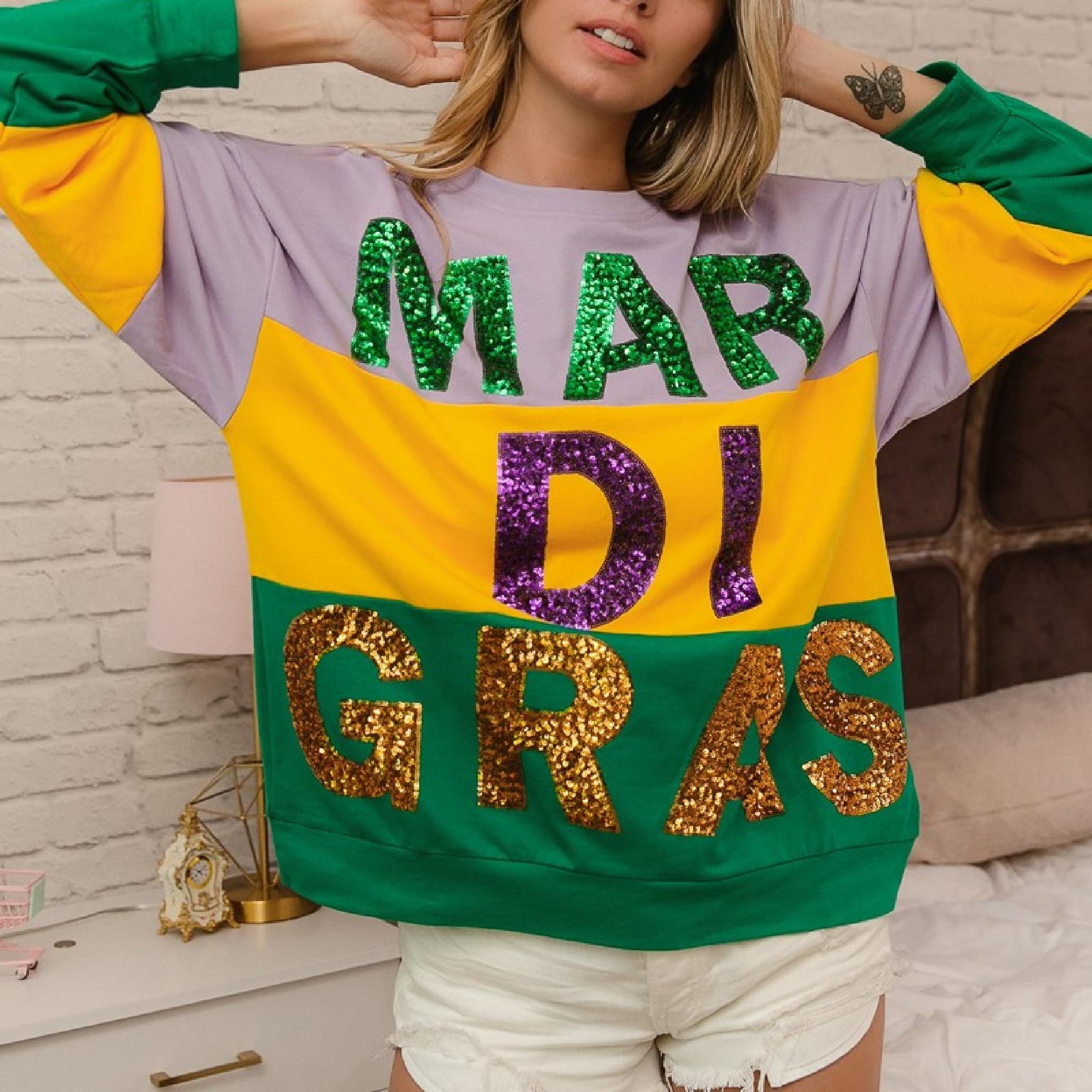 Person wearing a colorful 'Mardi Gras' sweatshirt with sequin letters against a brick wall.