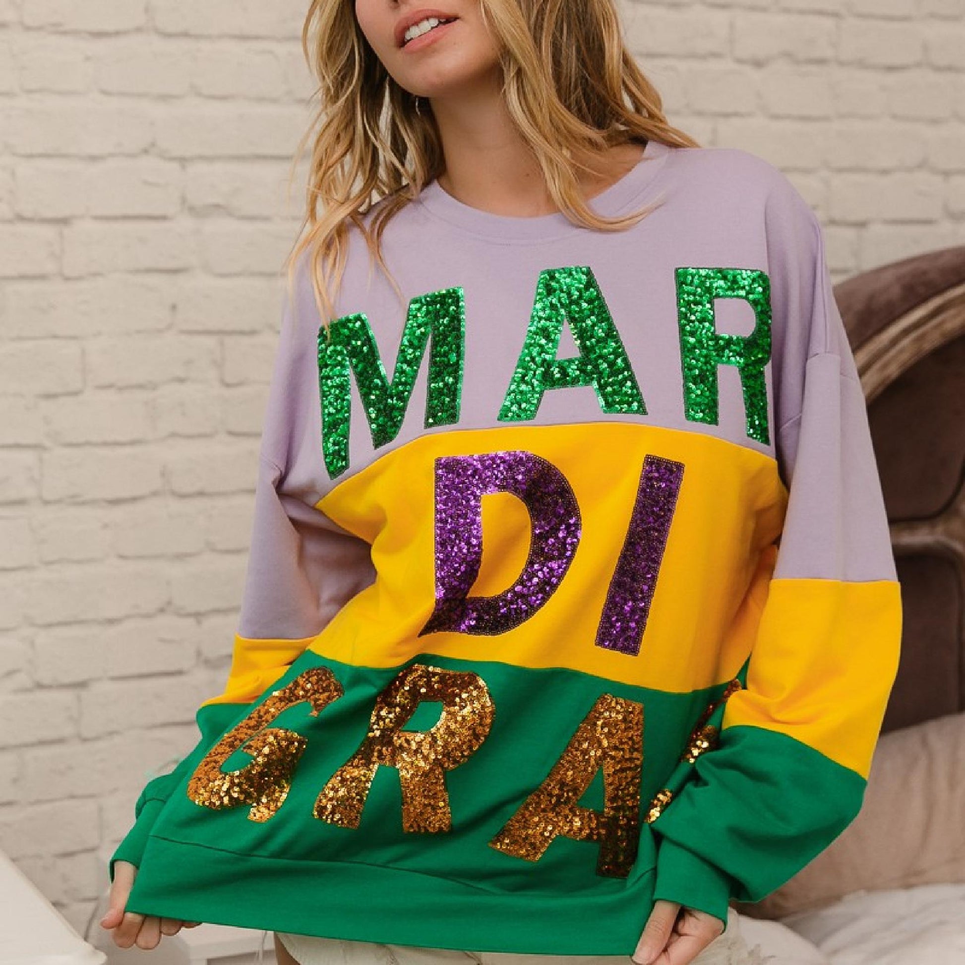 Person wearing a colorful 'Mardi Gras' sweatshirt with sequin letters against a brick wall.