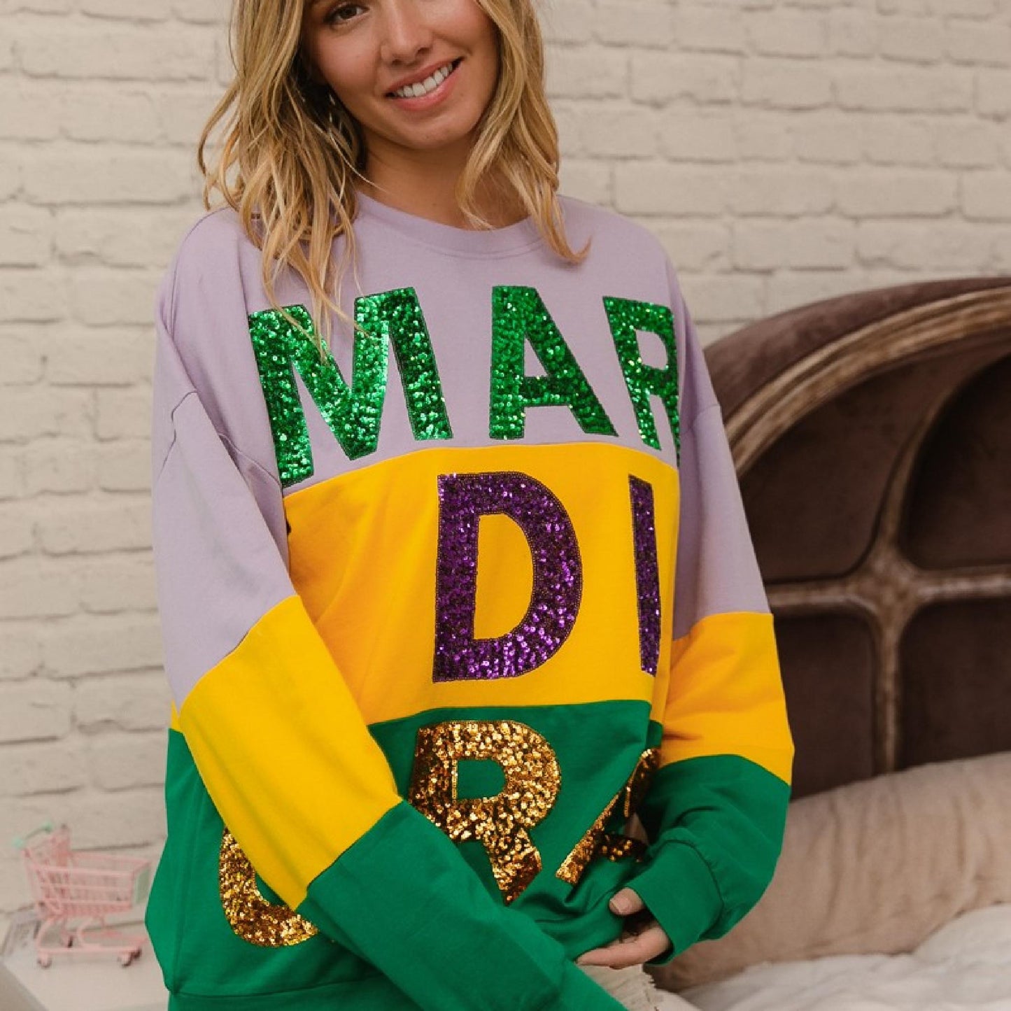 Person wearing a colorful sweatshirt with sequin letters in front of a brick wall.