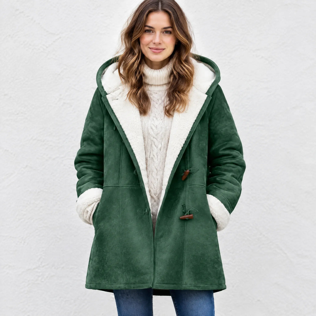 A woman wearing a dark green suede-like hooded coat with a cream sherpa lining, paired with a cream cable-knit turtleneck sweater and blue jeans, standing against a light textured wall.
