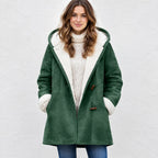 A woman wearing a dark green suede-like hooded coat with a cream sherpa lining, paired with a cream cable-knit turtleneck sweater and blue jeans, standing against a light textured wall.