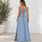 A woman wearing a light blue sleeveless maxi dress with a lace-up open back, shown from the back in an outdoor setting with a white wall and potted plant.