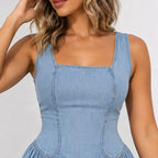 A close-up of a woman wearing a light blue denim corset-style dress with a square neckline, structured seams, and a gathered skirt, against a plain light gray background.