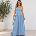 A woman modeling a sleeveless, light blue denim maxi dress with a structured corset-style bodice and gathered skirt, standing in front of a white wall next to a potted plant.