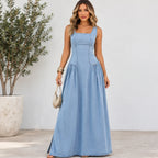 A woman modeling a sleeveless, light blue denim maxi dress with a structured corset-style bodice and gathered skirt, standing in front of a white wall next to a potted plant.