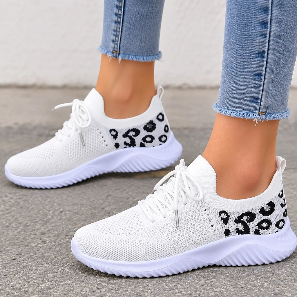 White slip-on knit sneakers with black leopard print details on the sides, shown on a model wearing light blue frayed-hem jeans.