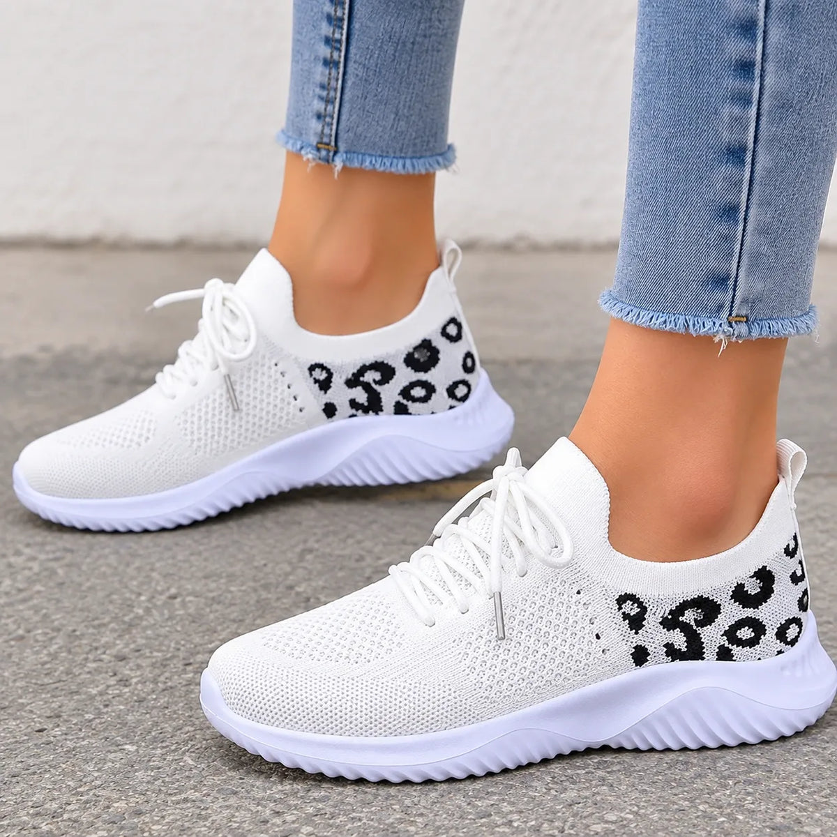 White slip-on knit sneakers with black leopard print details on the sides, shown on a model wearing light blue frayed-hem jeans.