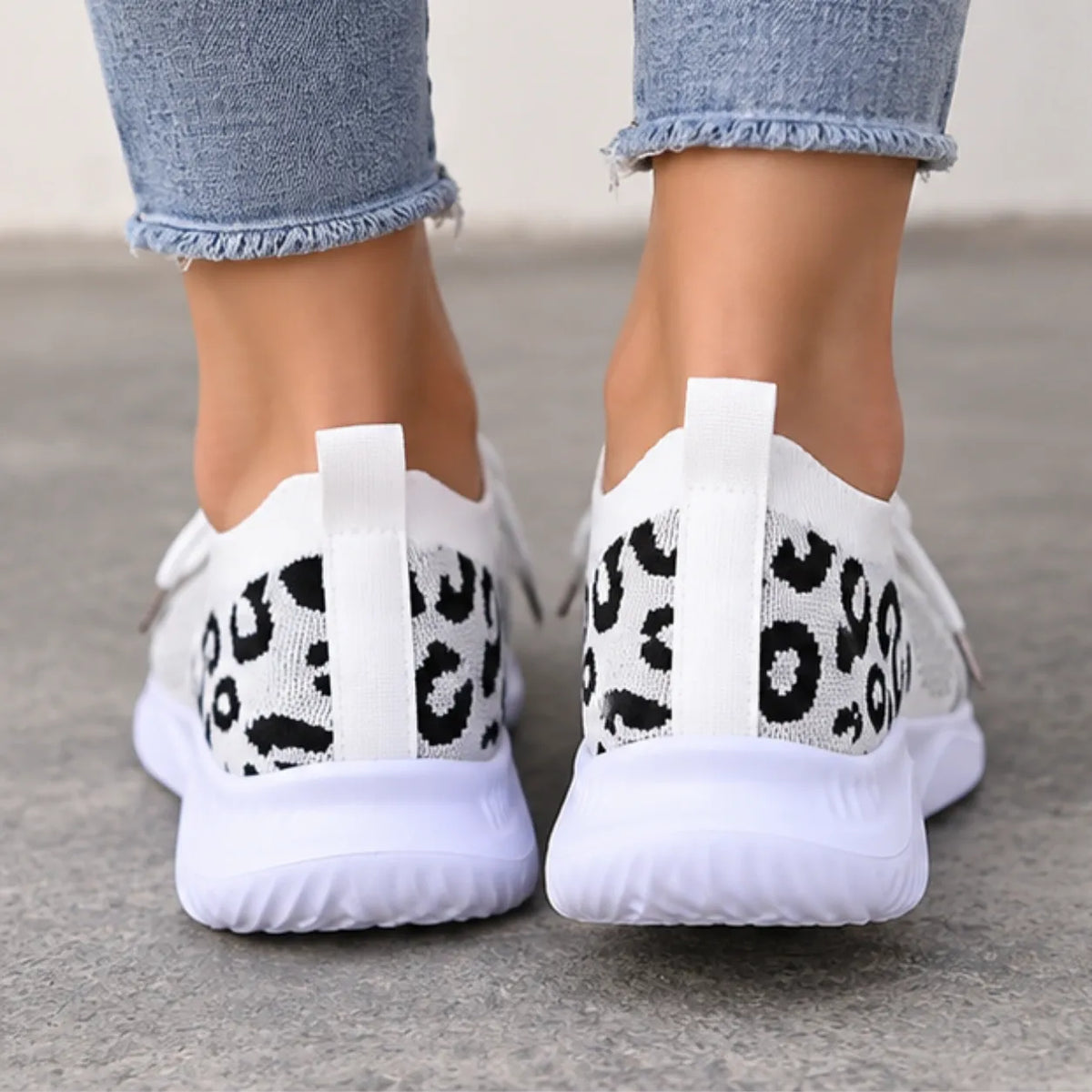 White slip-on knit sneakers with a black leopard print pattern on the heel, shown on a person wearing light blue frayed-hem jeans.