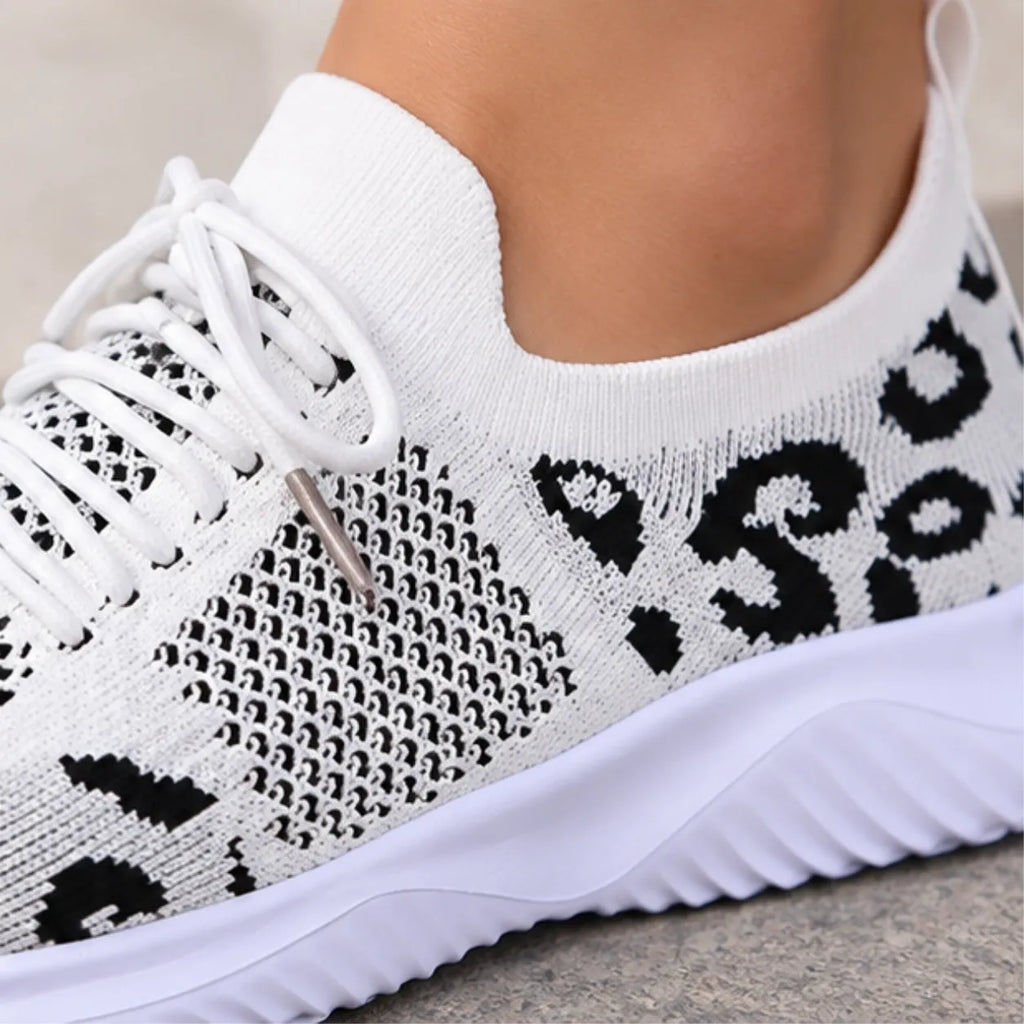A close-up of a white knit slip-on sneaker with black abstract pattern details and white laces, shown worn on a foot against a neutral outdoor background.