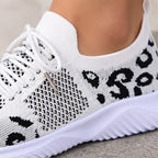 A close-up of a white knit slip-on sneaker with black abstract pattern details and white laces, shown worn on a foot against a neutral outdoor background.