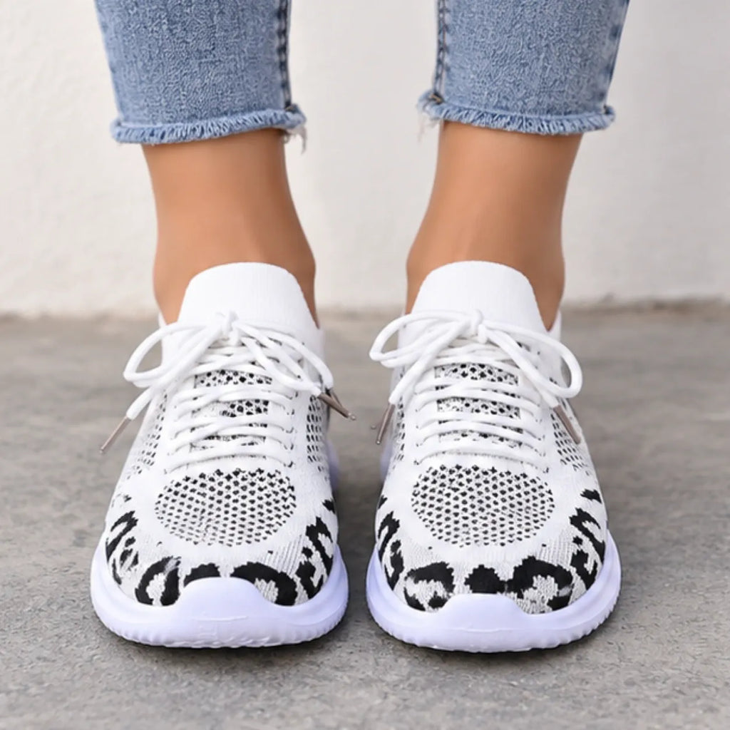 White knit sneakers with black leopard print detailing on the sides, shown on a model wearing light blue jeans against a neutral concrete background.