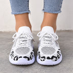 White knit sneakers with black leopard print detailing on the sides, shown on a model wearing light blue jeans against a neutral concrete background.