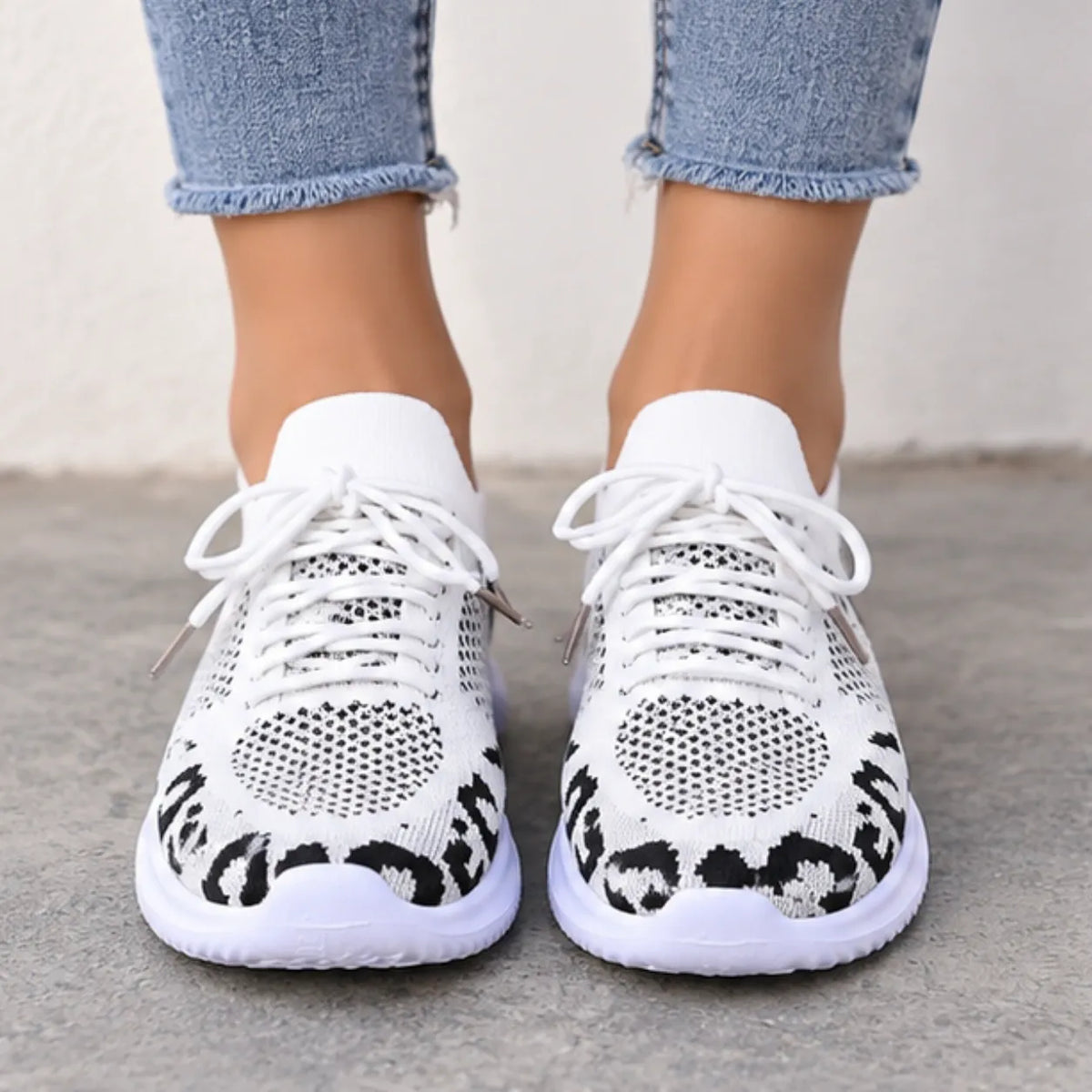 White knit sneakers with black leopard print detailing on the sides, shown on a model wearing light blue jeans against a neutral concrete background.