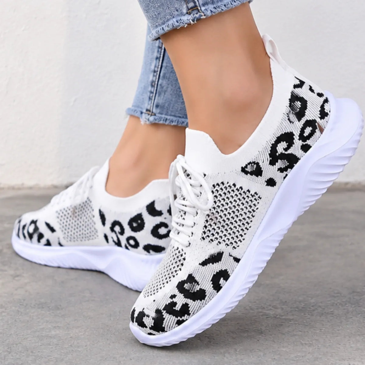 White knit slip-on sneakers with a black leopard print pattern, shown on a model wearing light blue jeans against a neutral concrete wall.