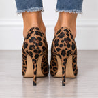 A pair of leopard print high-heeled pumps shown from the back, worn with frayed blue jeans on a light wood floor.