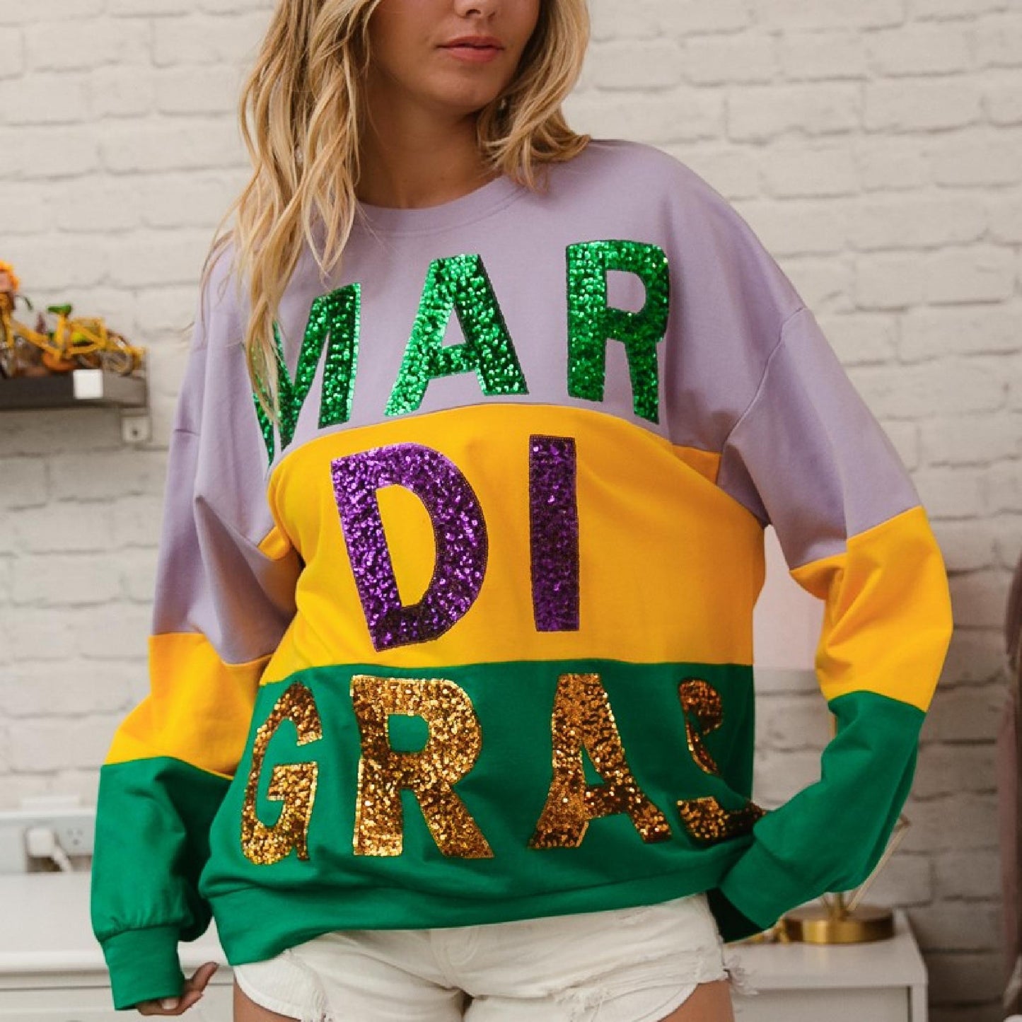 Person wearing a colorful 'MARDI GRAS' sweatshirt with sequin letters against a white brick wall.