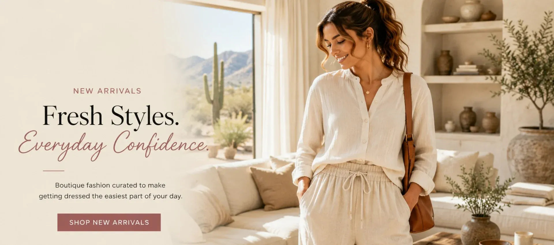 A woman stands in a bright, modern living room wearing a loose white long-sleeve blouse and a matching cream drawstring skirt. She has a brown shoulder bag and is posed in front of a window with desert scenery outside.