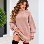 A woman wearing an oversized ribbed knit turtleneck sweater in a dusty rose pink color, styled with black thigh-high socks, standing in front of a modern white wall with a green plant on the left.