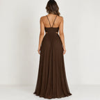 A woman viewed from the back wearing a floor-length brown dress with thin back straps and a pleated skirt, photographed against a plain light background.