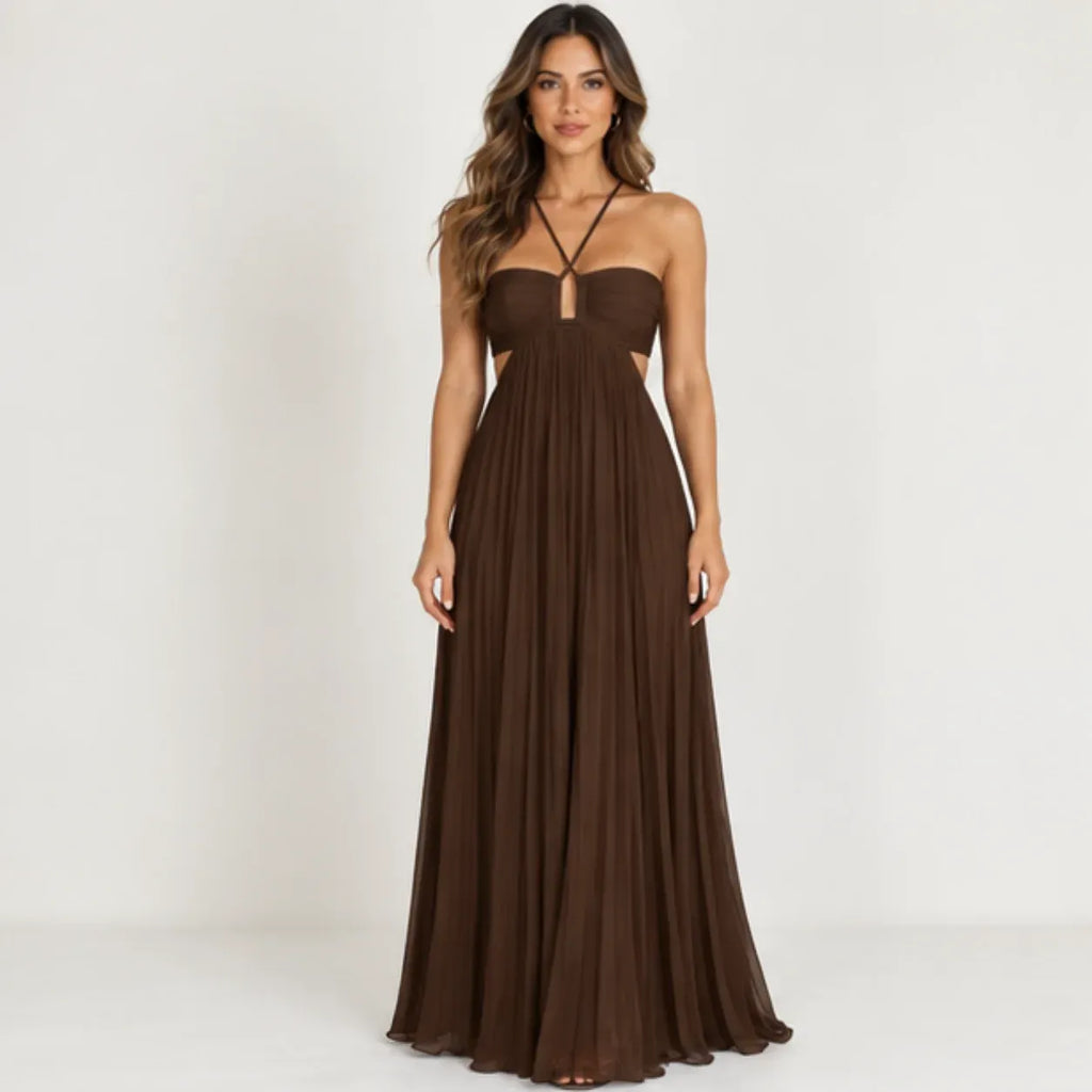 A woman modeling a floor-length brown strapless gown with a sweetheart neckline, thin crisscross halter straps, side cutouts, and a flowing pleated skirt against a plain light background.