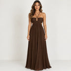 A woman modeling a floor-length brown strapless gown with a sweetheart neckline, thin crisscross halter straps, side cutouts, and a flowing pleated skirt against a plain light background.