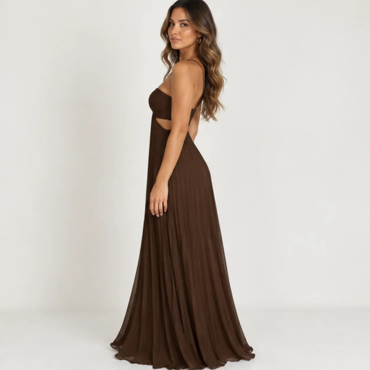 A woman modeling a floor-length brown strapless gown with side cutouts and a flowing pleated skirt, photographed against a plain light gray studio background.