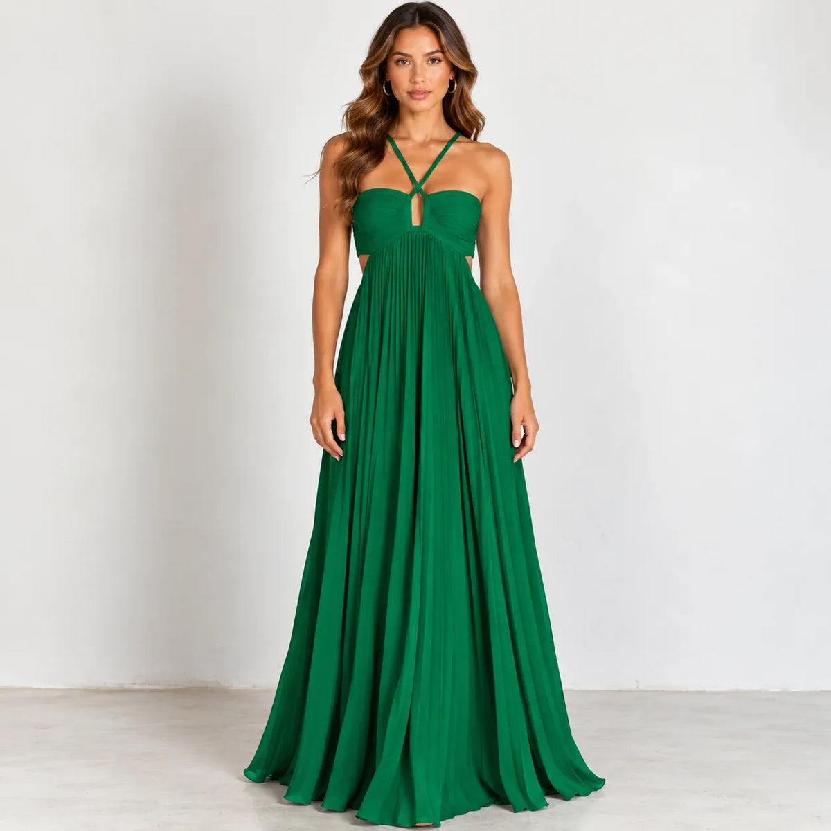 A woman modeling a floor-length emerald green halter gown with a sweetheart neckline, keyhole cutout, and side cutouts, standing against a plain light gray studio wall.