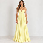 A woman modeling a pale yellow strapless maxi dress with a sweetheart neckline, thin crossover halter straps, side cutouts, and a flowing pleated skirt against a plain light gray studio background.