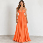 A woman modeling a bright orange strapless maxi dress with a sweetheart neckline, thin crossover straps, and a flowing pleated skirt, standing against a plain off-white wall.