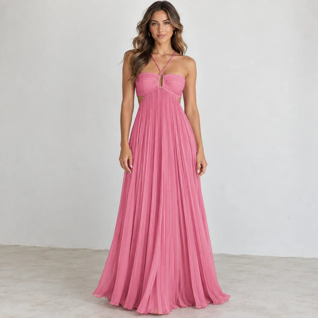 A woman modeling a floor-length pink halter gown with a pleated skirt, photographed against a plain light gray studio wall and floor.
