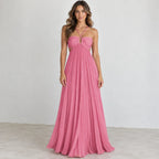 A woman modeling a floor-length pink halter gown with a pleated skirt, photographed against a plain light gray studio wall and floor.