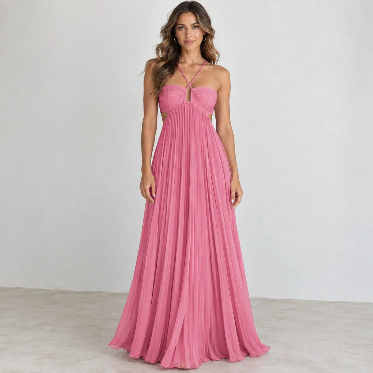 A woman modeling a floor-length pink halter gown with a pleated skirt, photographed against a plain light gray studio wall and floor.
