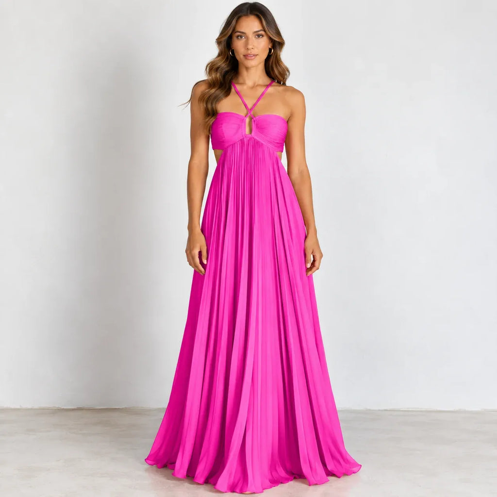 A woman modeling a bright hot pink halter-neck maxi dress with a sweetheart neckline, pleated bodice, and flowing skirt, standing against a plain light gray studio background.
