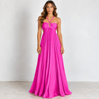 A woman modeling a bright hot pink halter-neck maxi dress with a sweetheart neckline, pleated bodice, and flowing skirt, standing against a plain light gray studio background.