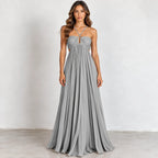 A woman modeling a floor-length gray halter gown with a ruched sweetheart bodice, side cutouts, and a flowing pleated skirt, standing against a plain light gray wall.