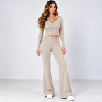 A woman modeling a matching beige two-piece lounge set consisting of a cropped zip-up hoodie with long sleeves and high-waisted flared pants, paired with white sneakers, against a light gray studio background.