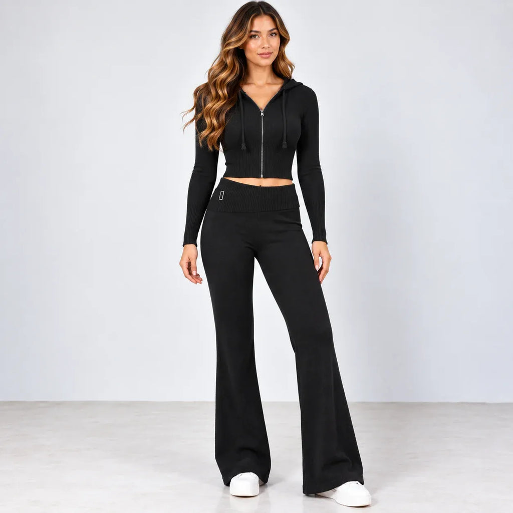A woman modeling a black two-piece activewear set consisting of a cropped zip-up hoodie with long sleeves and matching high-waisted flare pants, paired with white sneakers against a light gray studio background.