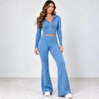 A woman modeling a matching light blue two-piece loungewear set consisting of a cropped zip-up hoodie and high-waisted flare pants, paired with white sneakers against a neutral studio background.