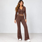 A woman modeling a matching brown knit two-piece set consisting of a cropped zip-front hoodie top and high-waisted flared pants, paired with white sneakers against a light gray studio background.