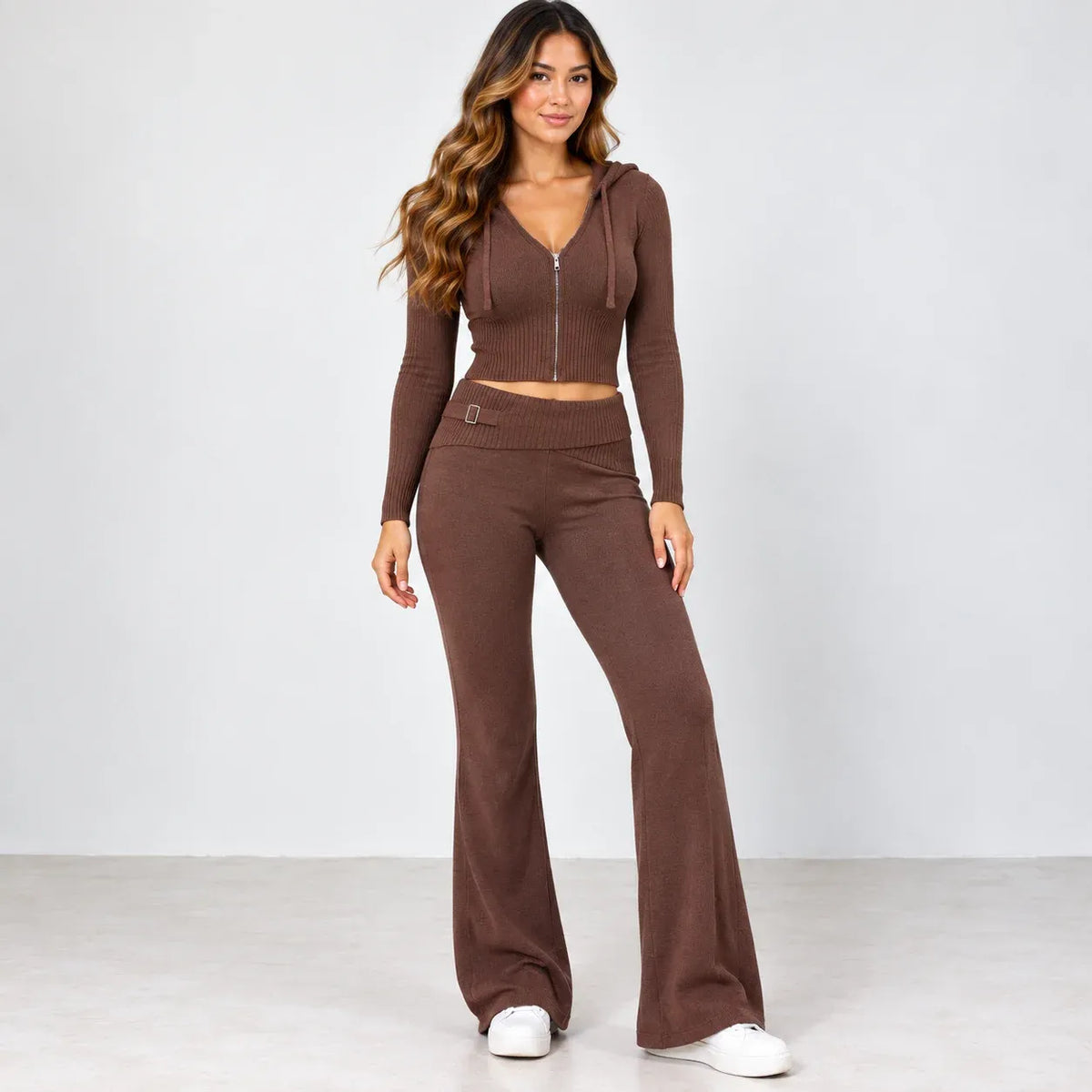A woman modeling a matching brown knit two-piece set consisting of a cropped zip-front hoodie top and high-waisted flared pants, paired with white sneakers against a light gray studio background.