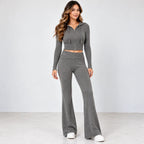 A woman modeling a matching gray two-piece knit loungewear set, featuring a cropped zip-up hoodie with drawstrings and high-waisted, wide-leg flare pants, paired with white sneakers against a light gray studio background.